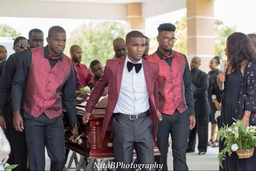 Pallbearers in burgundy vests and black bow ties carry a coffin at an outdoor funeral ceremony.
