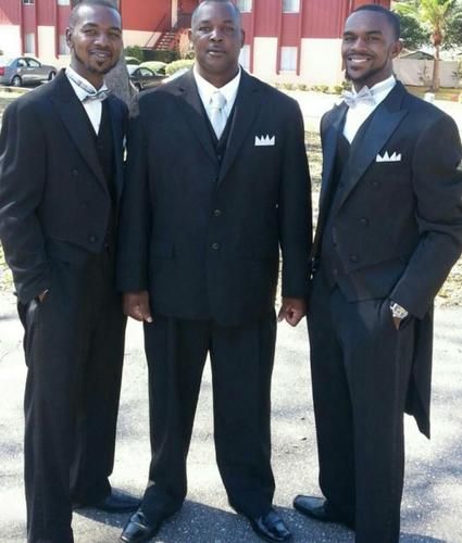 Three men in black suits standing together outdoors, smiling at the camera.