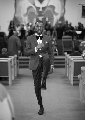 Man in a dark suit walking down a church aisle toward the camera, with people seated in the background.