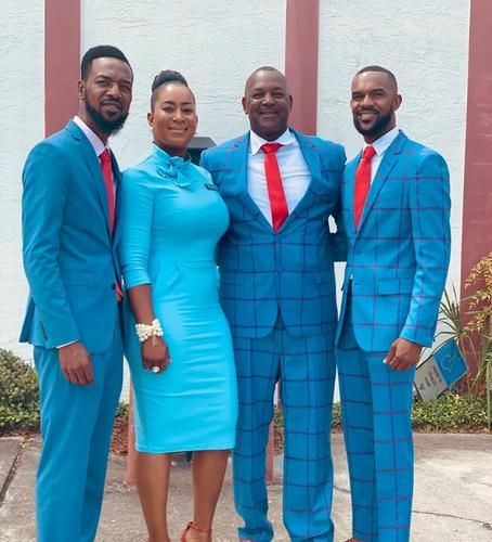 Four people in matching blue outfits and red ties posing outdoors against a white wall.