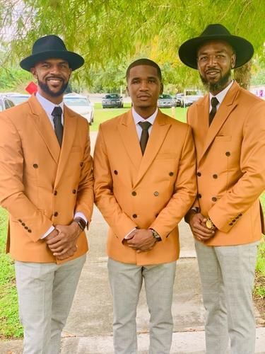 Three men in orange blazers and gray pants stand outdoors, smiling beneath trees.