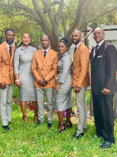 Group of six people posing outdoors on grass, four in tan blazers and two in formal wear.