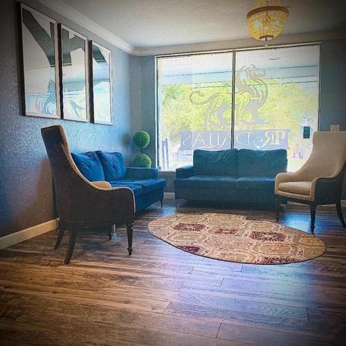 Bright lounge with blue sofas, tan chairs, round rug, and large sunlit window.