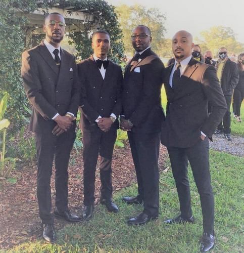 Four people in black suits stand outdoors in a garden at a formal event.
