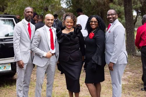 Five people in formal black, gray, and red outfits posing outdoors at a gathering.