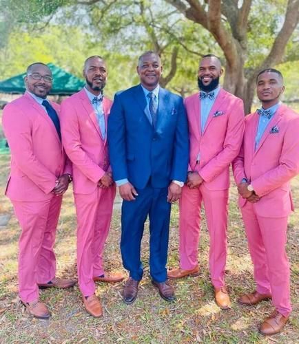 Five men in colorful suits pose outdoors beneath trees, with one in a blue suit centered among four in pink.