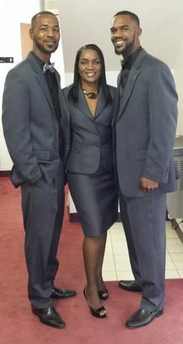 Three people in gray suits posing together on a red carpet indoors, smiling at the camera.
