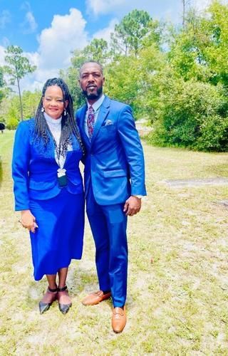 Two people in blue outfits standing together outdoors on a grassy path with trees in the background