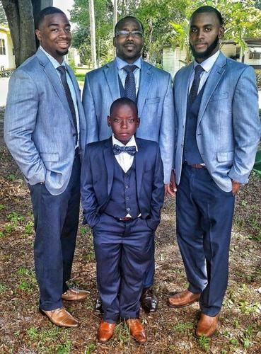 Four people in blue suits posing outdoors under trees, with one child in front.
