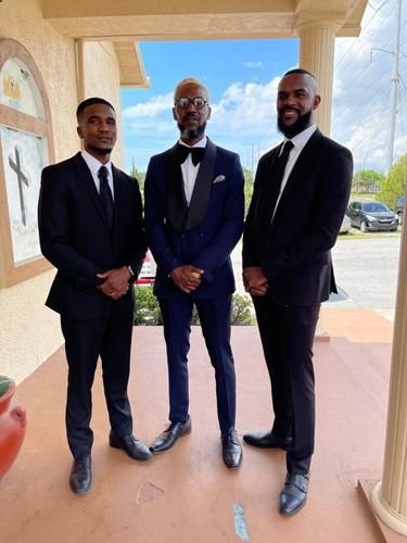 Three men in dark suits pose outdoors by a porch with columns and a bright sky.