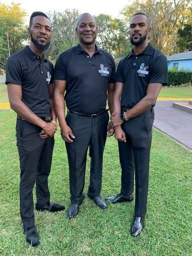 Three men in black polo shirts and pants standing on grass outdoors, posing in front of a road.