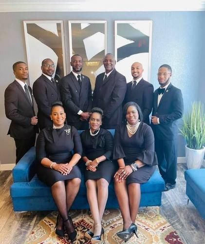 Group of people in formal black attire posing in an office lounge with a blue couch and wall art.
