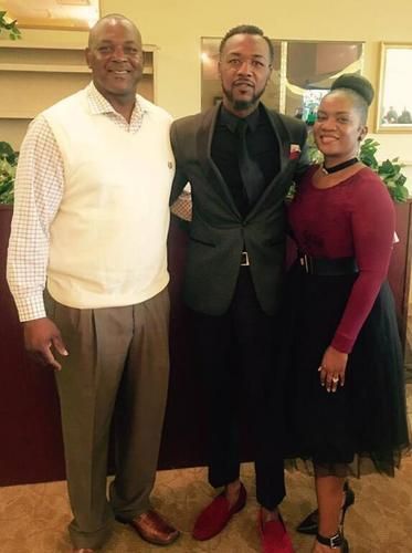 Three people posing indoors, with one man in a black suit and red shoes centered between two others.