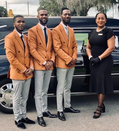 Four people standing beside a black limousine, wearing coordinated orange blazers and formal black outfits.