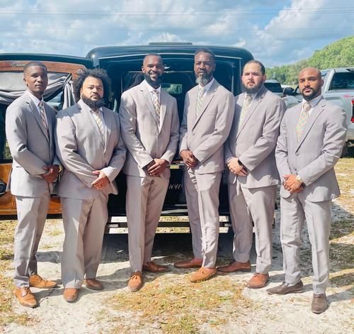 Six men in light gray suits posing beside a black SUV under a bright sky