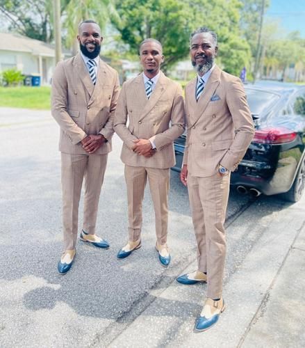 Three men in tan suits pose on a sidewalk beside a black car on a sunny street.