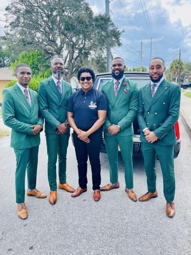Five men in matching green suits pose outdoors on a road, with one in black standing in front.