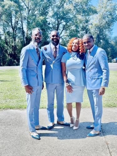 Four people in light blue suits posing outdoors on a sunny path by trees