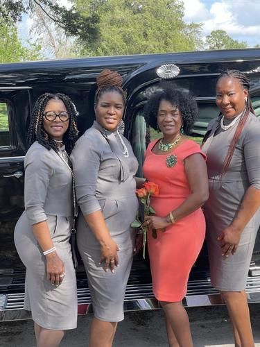 Four women pose in front of a black van, wearing matching gray dresses and one coral dress.
