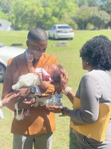 Two adults outdoors holding a baby, with a car and green field in the background.