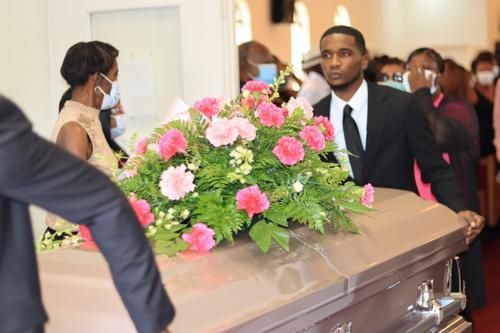 Casket with pink and white floral arrangement at a funeral, with mourners gathered around it