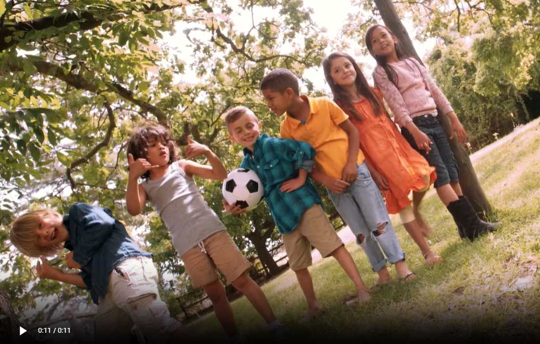 Children playing with a soccer ball in a grassy park, smiling under trees