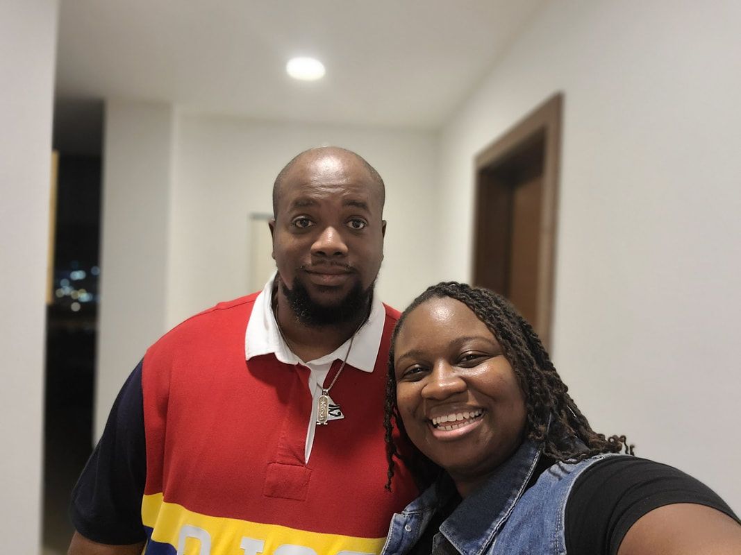 Two people smiling indoors in a hallway, one in a red shirt and one in a black top.