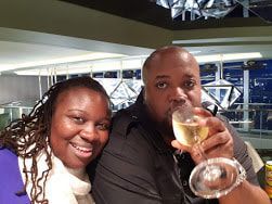 Two people smiling at a table, one holding a glass of white wine in a bright indoor setting