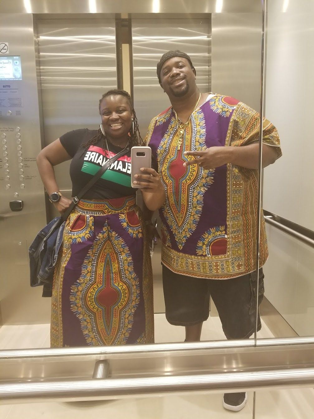 Two people smiling in an elevator, taking a mirror selfie in colorful patterned outfits.