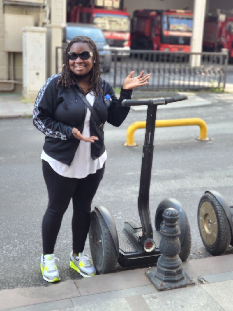 Woman standing on a sidewalk beside a scooter, smiling and waving, with a street and railing in the background