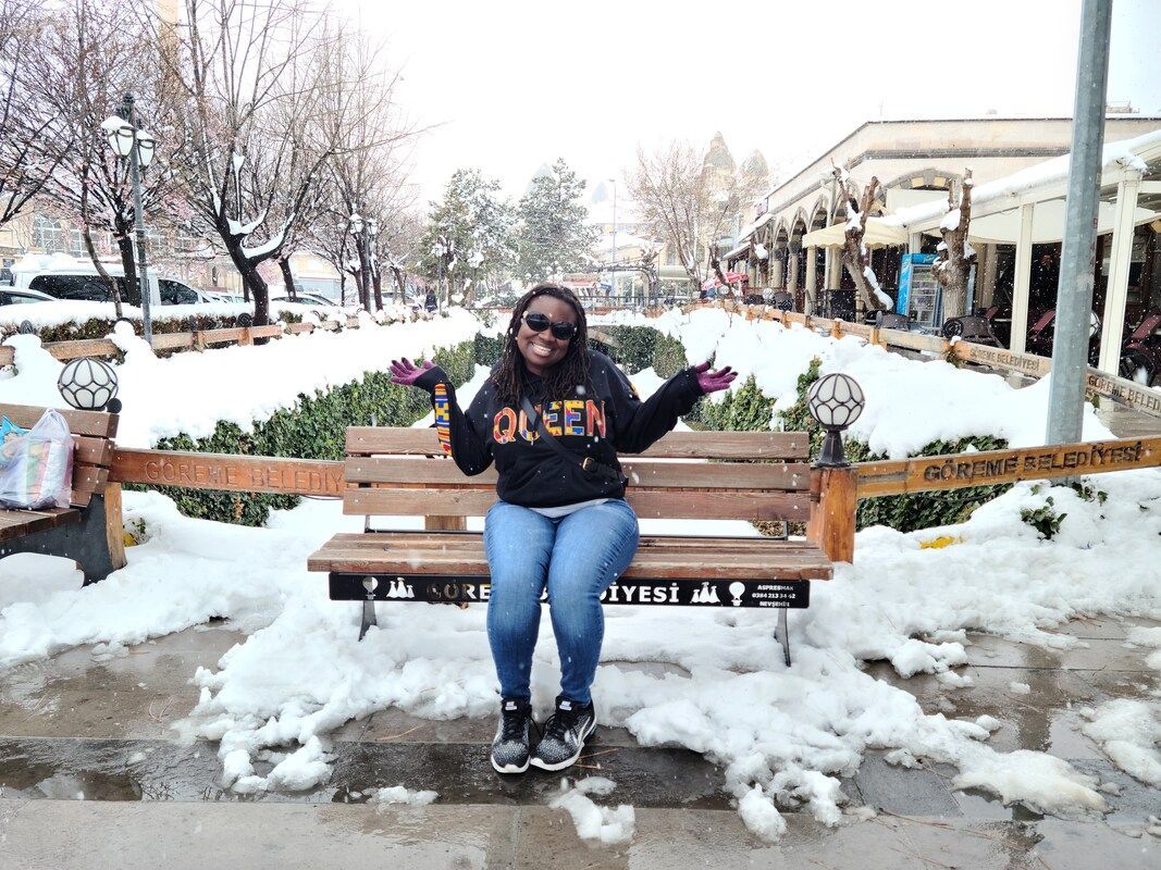 Person seated on a park bench in snowy garden, wearing sunglasses and a dark sweater with blue jeans.
