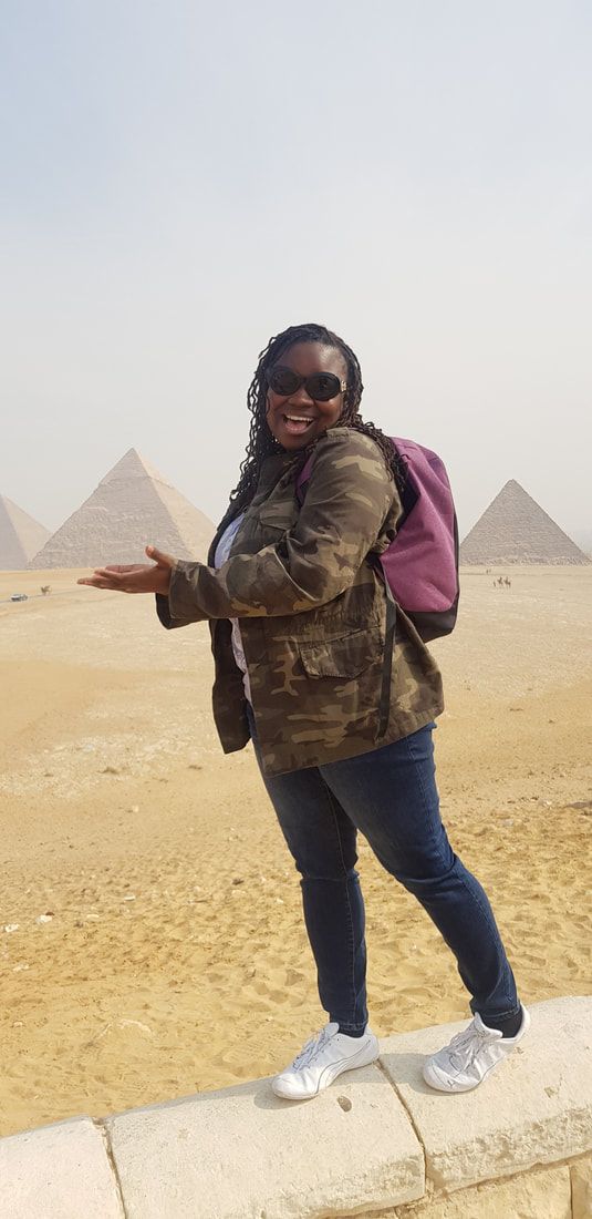 Person in sunglasses posing on a sandy plain with pyramids in the background.