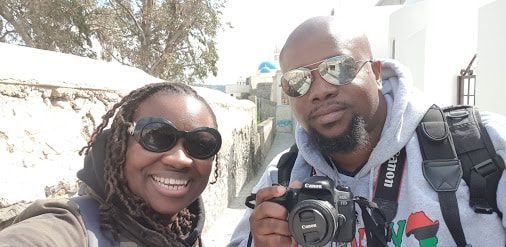 Two smiling people wearing sunglasses pose outdoors, one holding a camera.