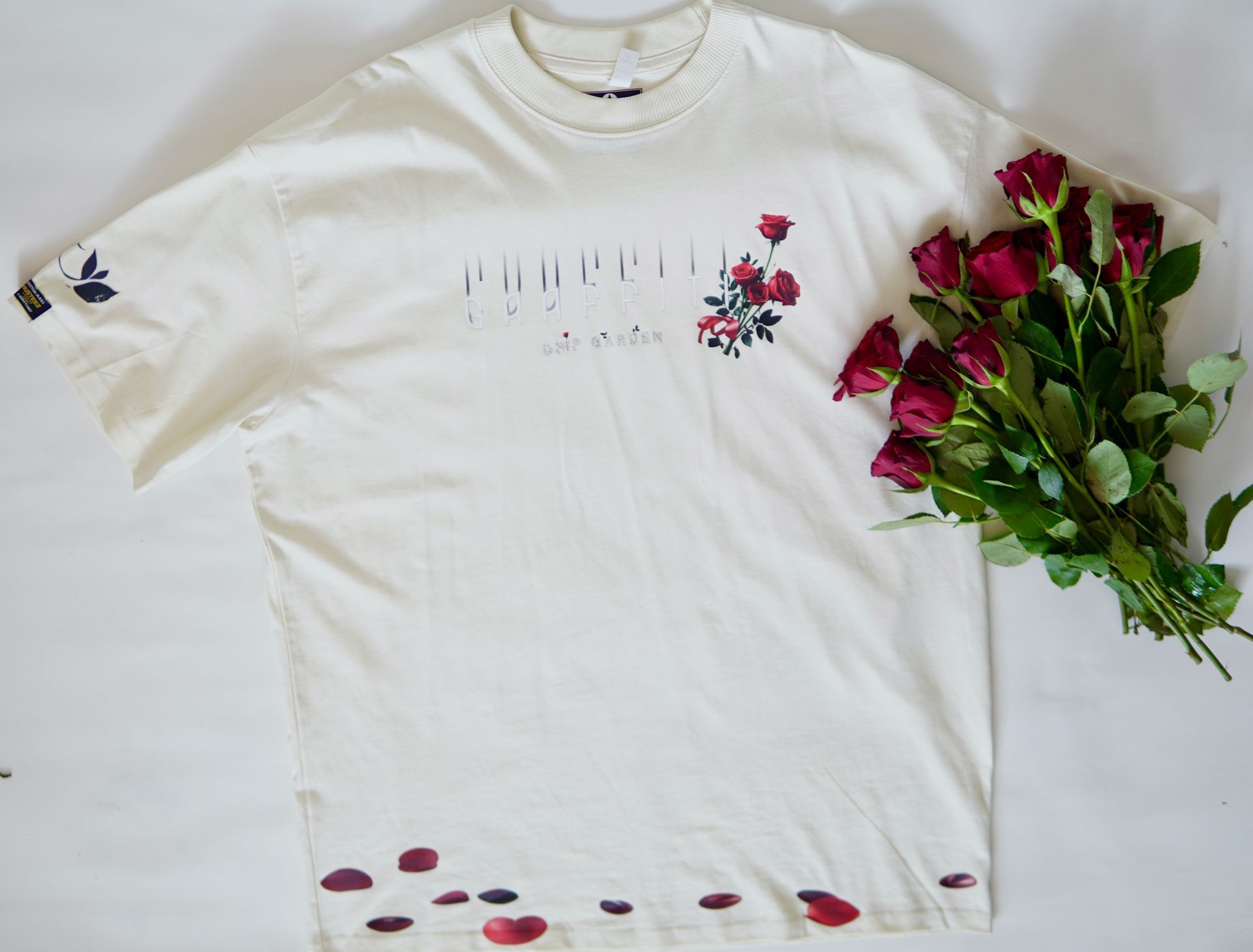 Cream-colored t-shirt with floral design, next to a bouquet of red roses and scattered petals.