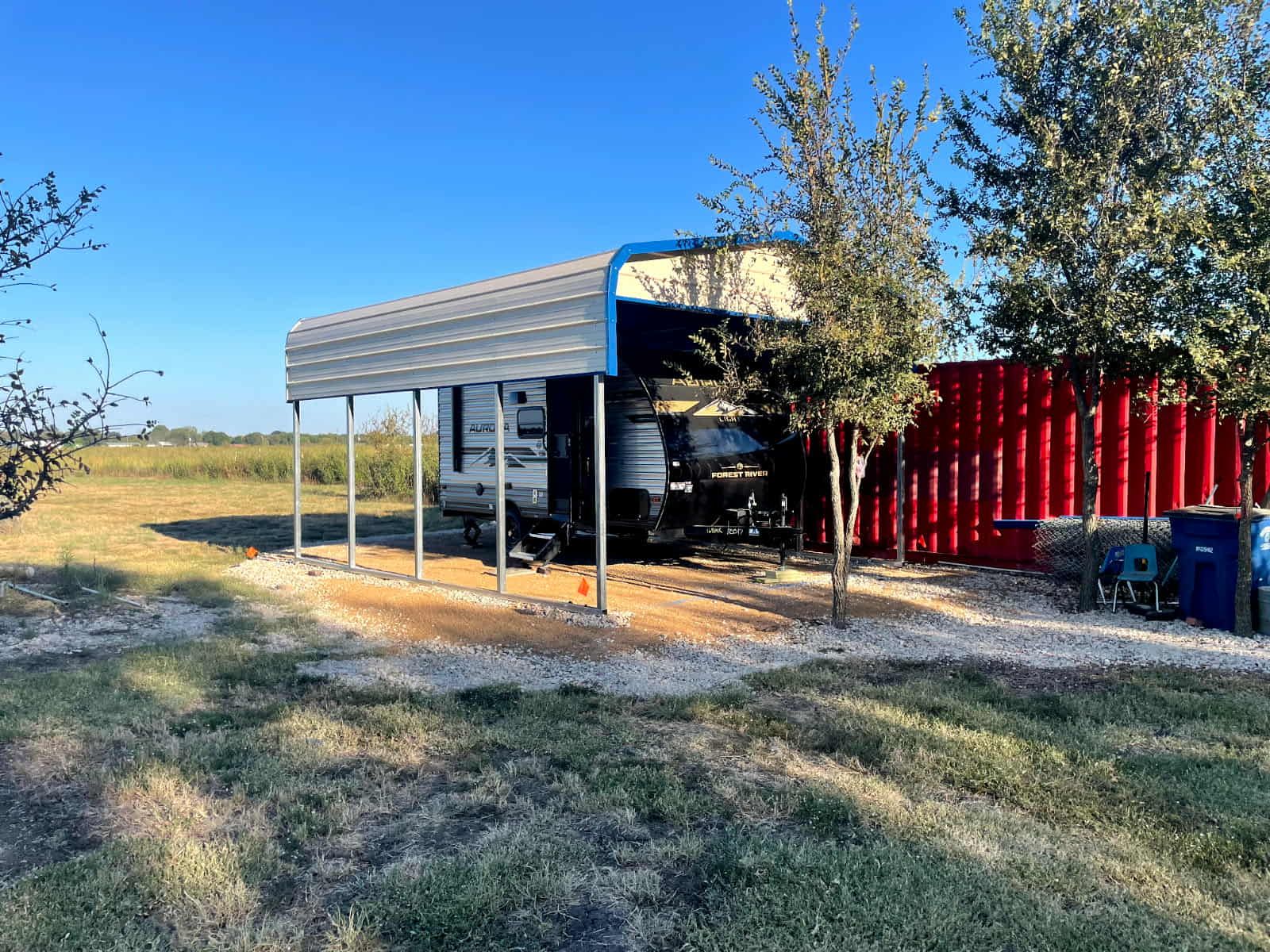 RV parked beside a small metal shed in a grassy rural field, with trees and red fence panels nearby