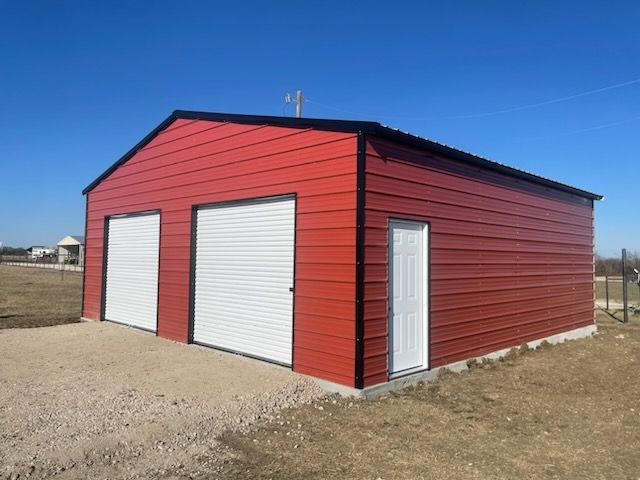 Red metal storage building with white roll-up doors on a gravel lot under a clear blue sky