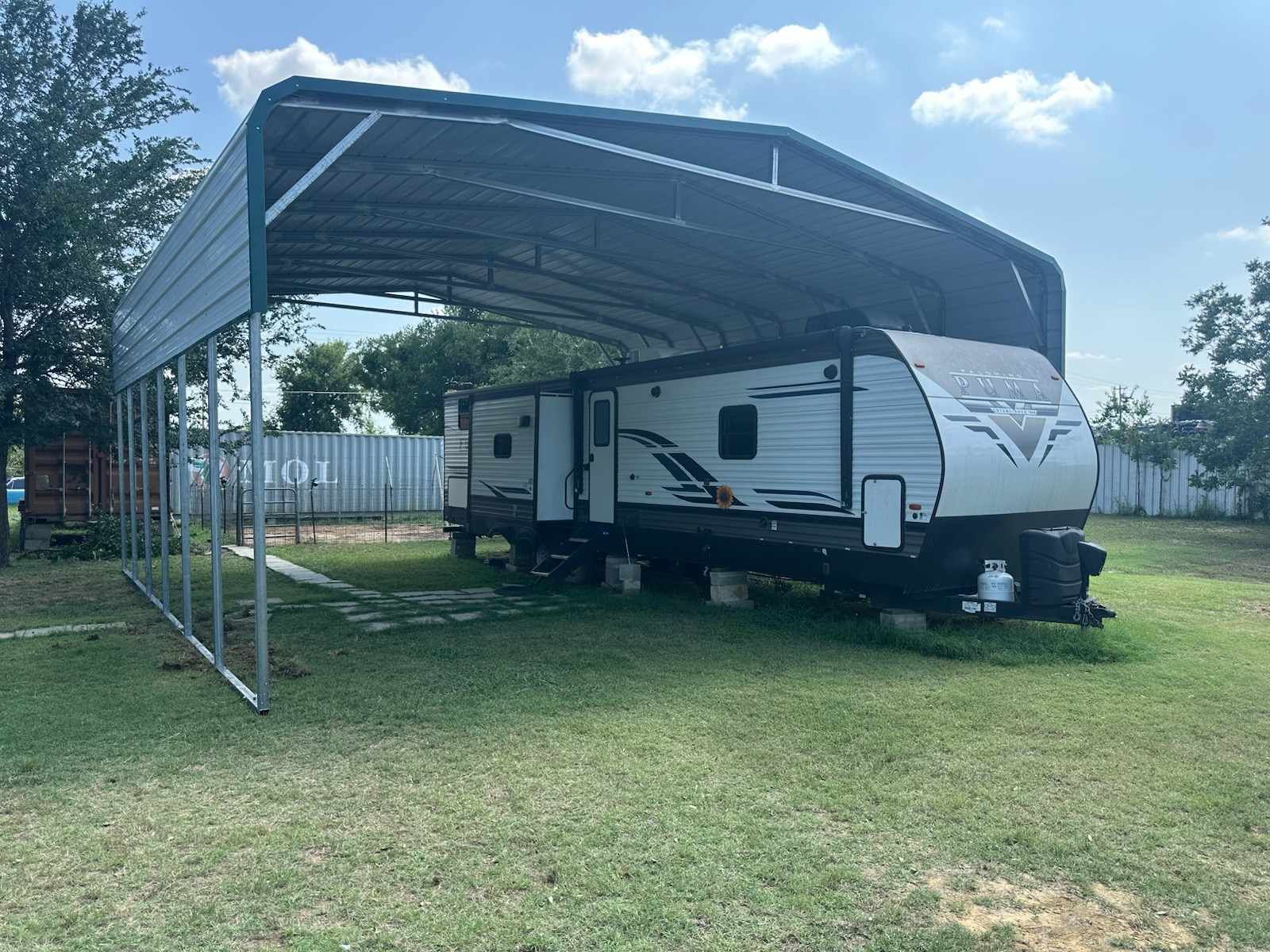 White and gray travel trailer parked under a metal carport on a grassy lot