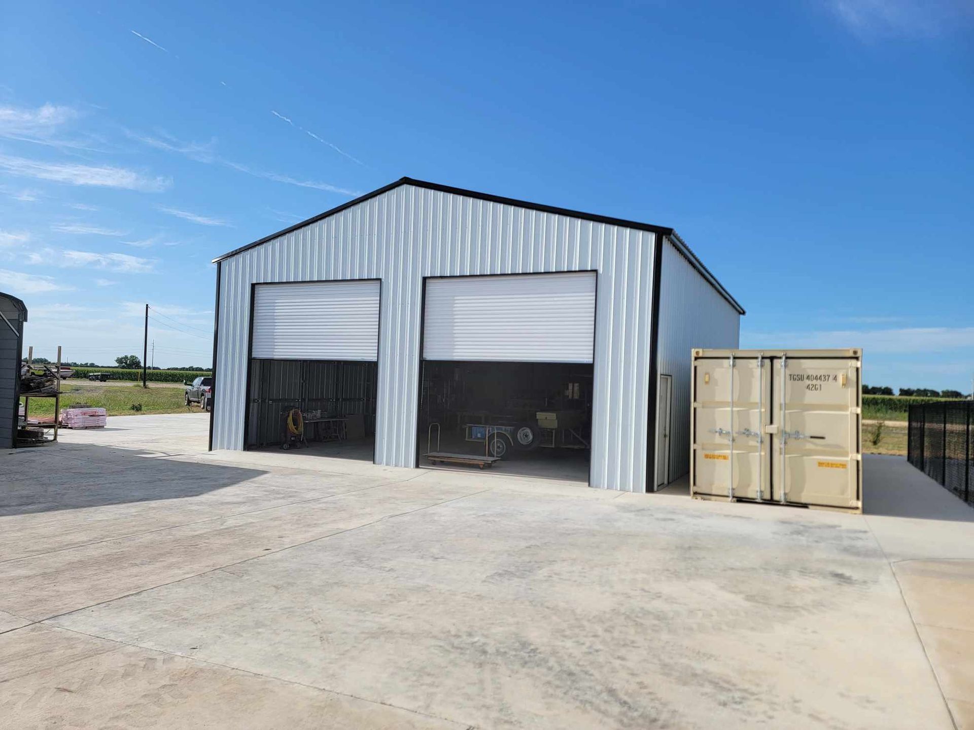 Metal warehouse with open bay doors and a beige shipping container on a concrete lot under a blue sky