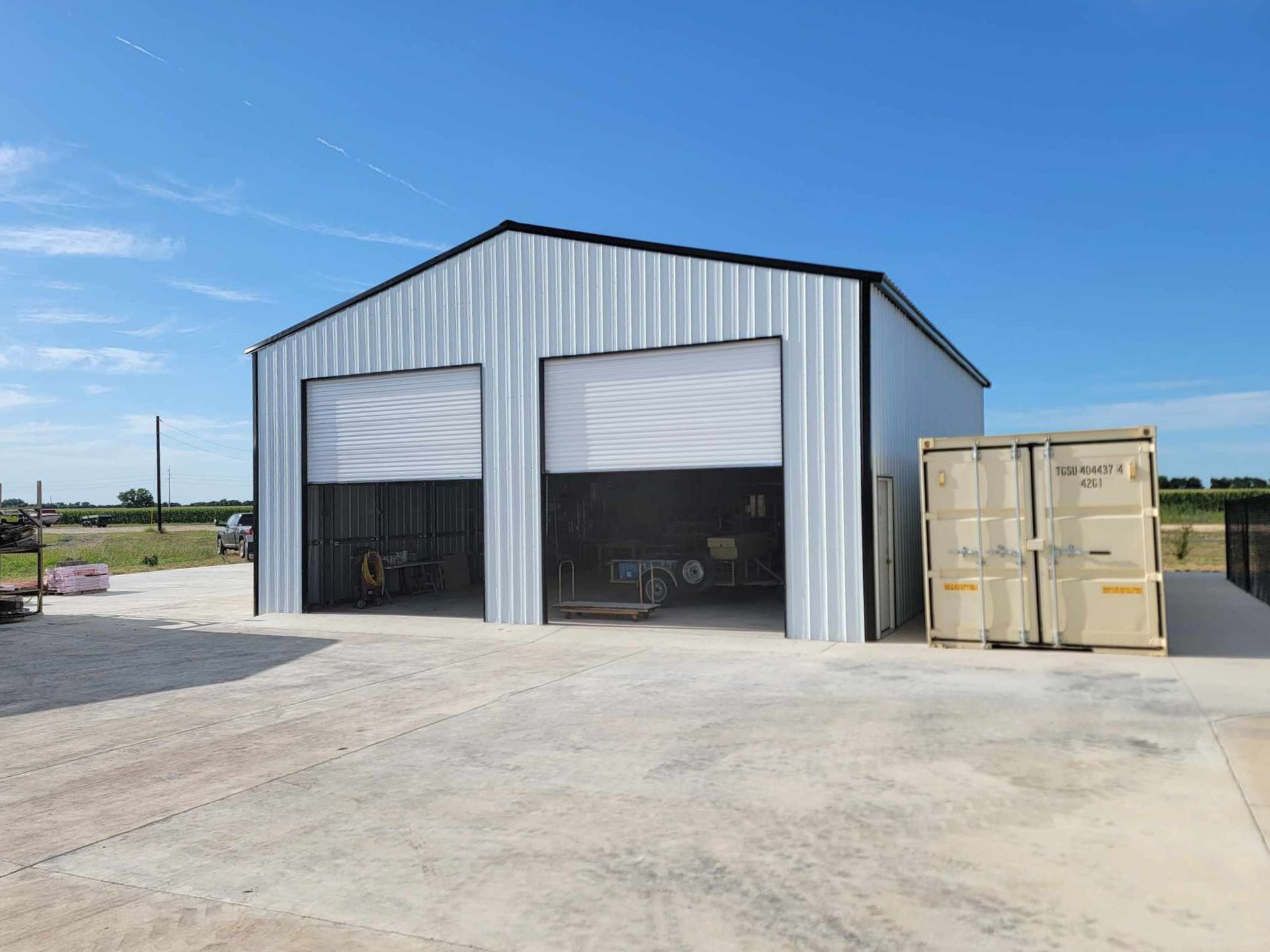 Metal warehouse with two open bays beside a beige shipping container on a paved lot under a blue sky