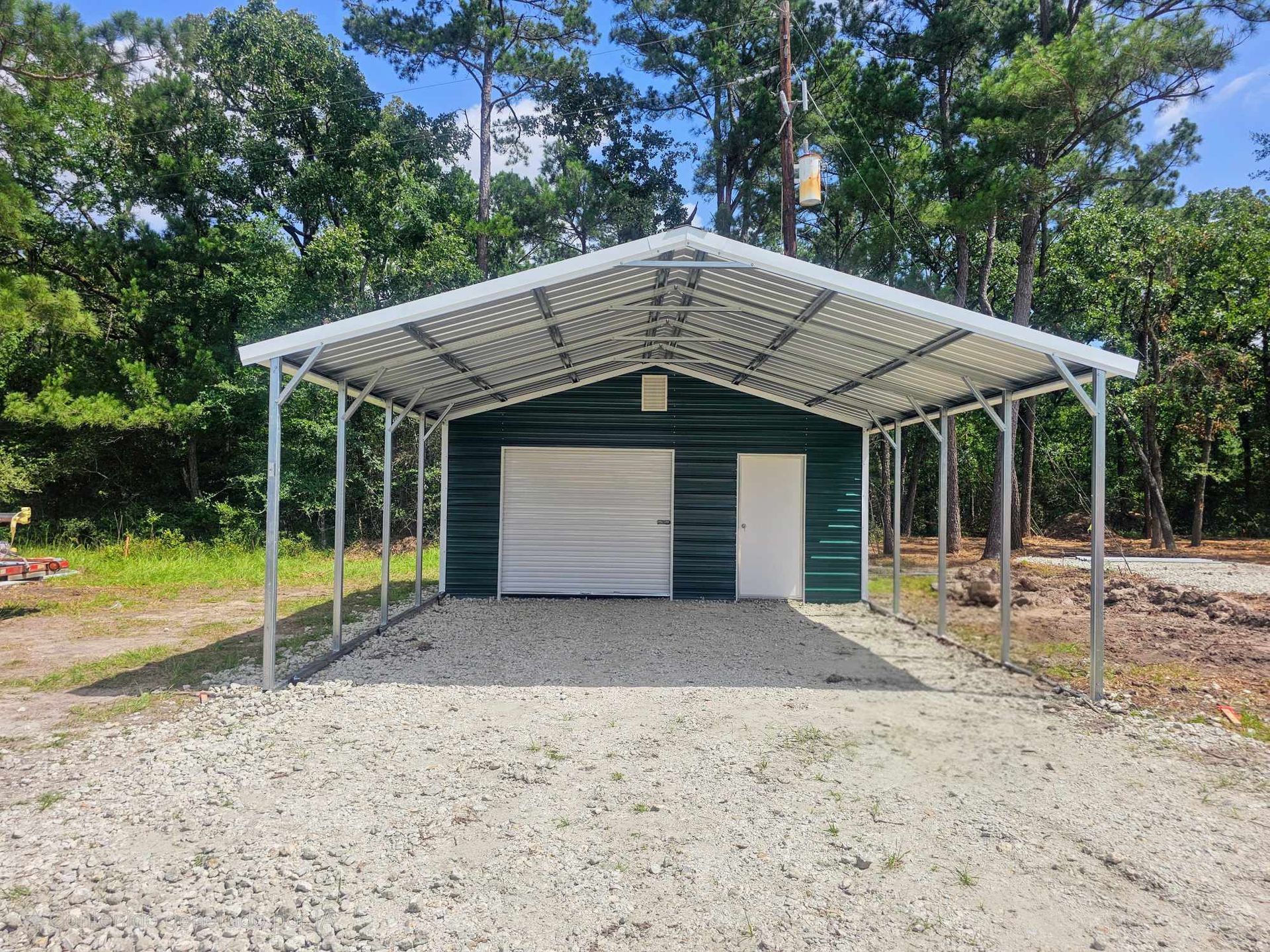 Covered metal carport shelter with two garage doors on a gravel lot, surrounded by trees.