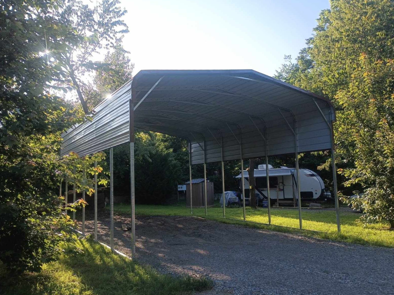 Metal carport beside a gravel drive, with a white van parked underneath in a wooded yard.