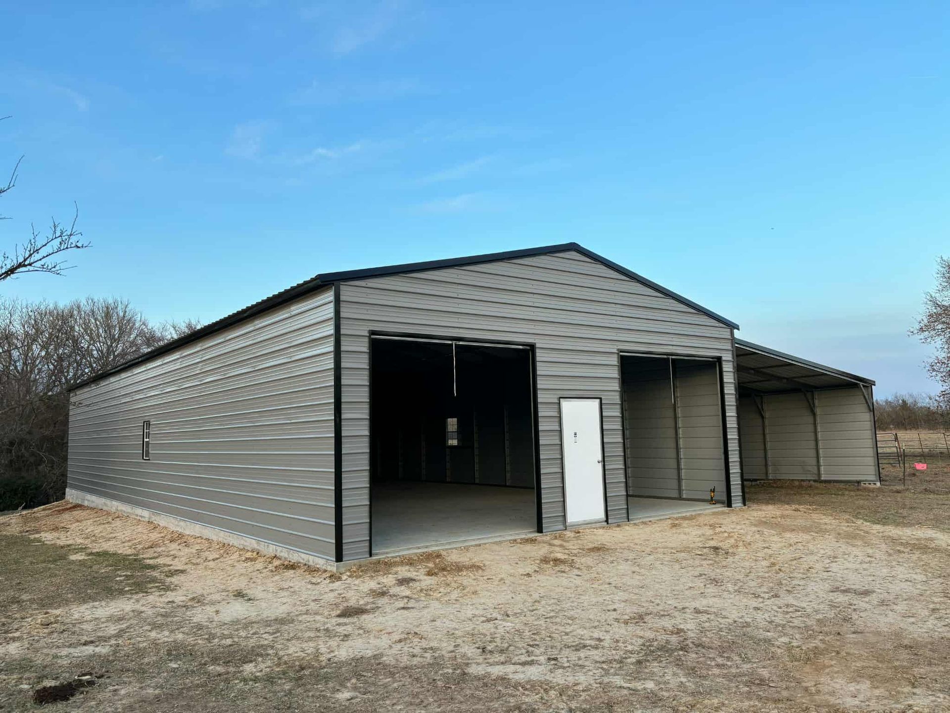 Gray metal garage building with open bays on a gravel lot under a blue sky