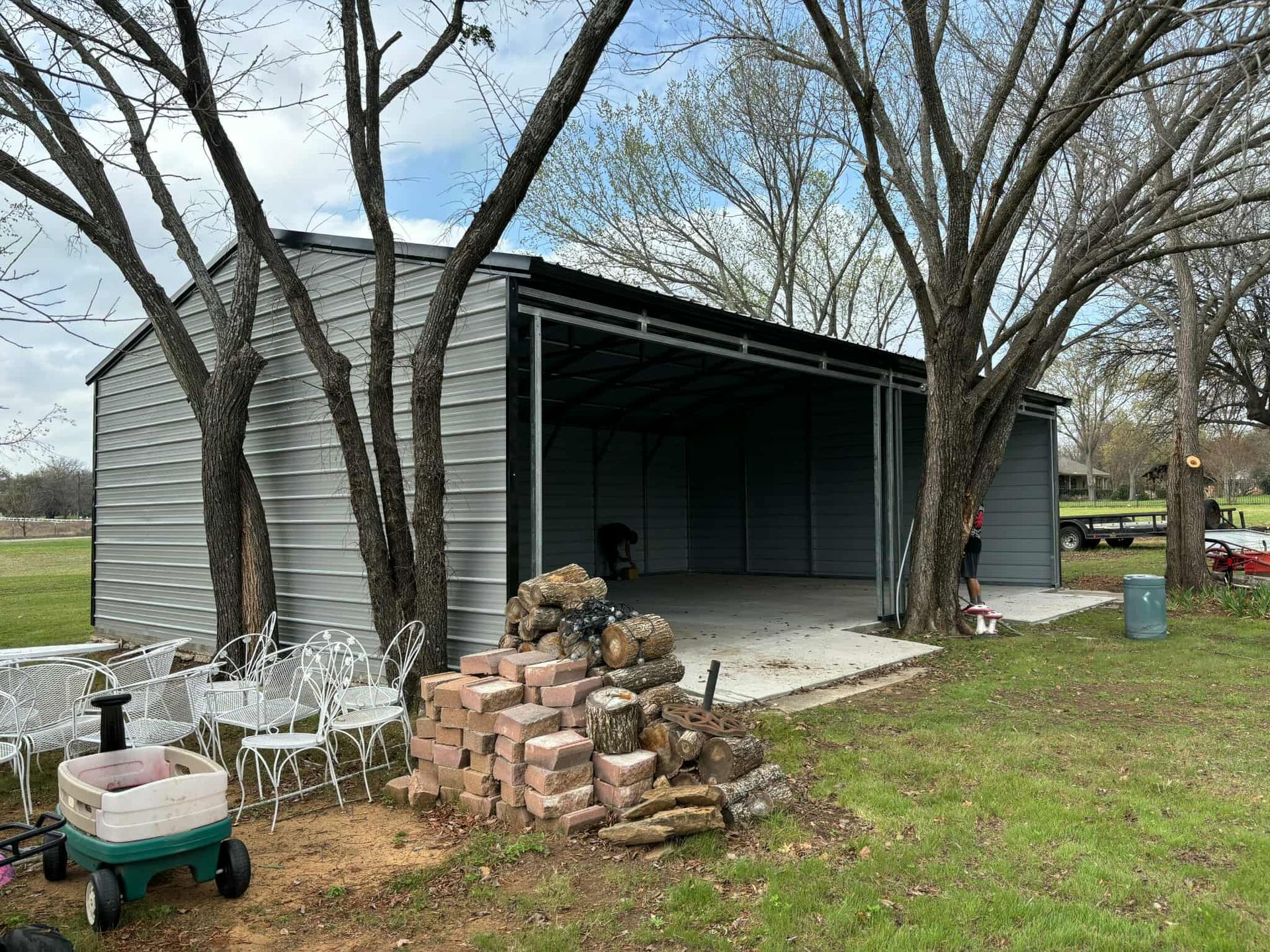 Open-sided metal shed with stacked bricks, trees, and yard items in a grassy yard