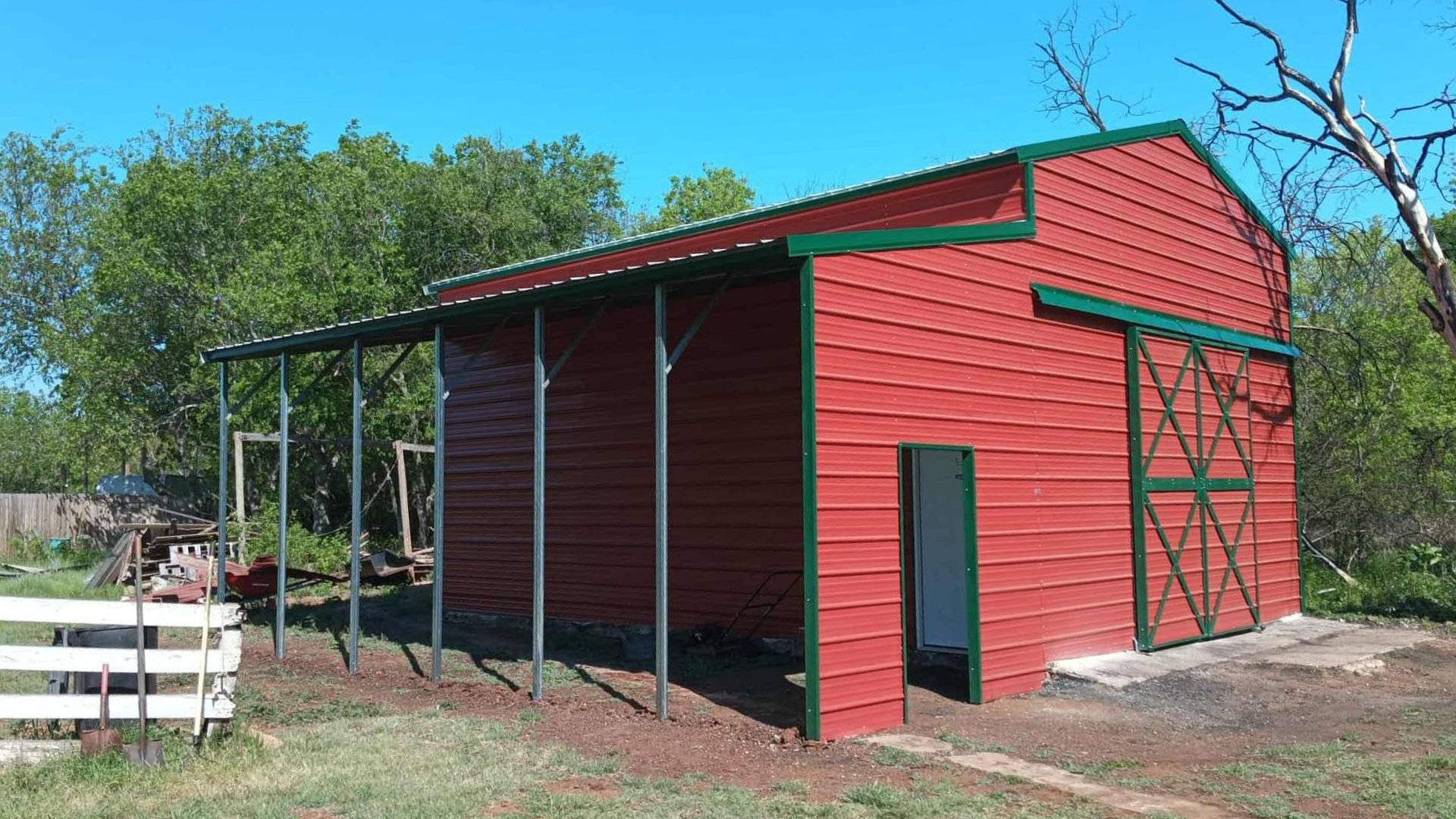 Red barn-style shed with green trim and covered side porch in a grassy rural yard under a blue sky