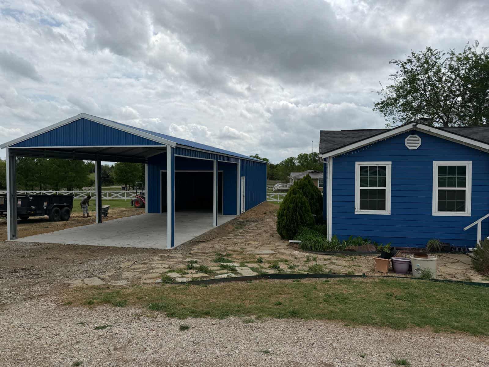 Blue house and  metal carport on a gravel lot under a cloudy sky.