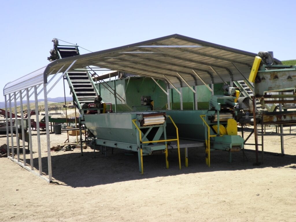 Large green industrial machine under a metal canopy in an outdoor yard