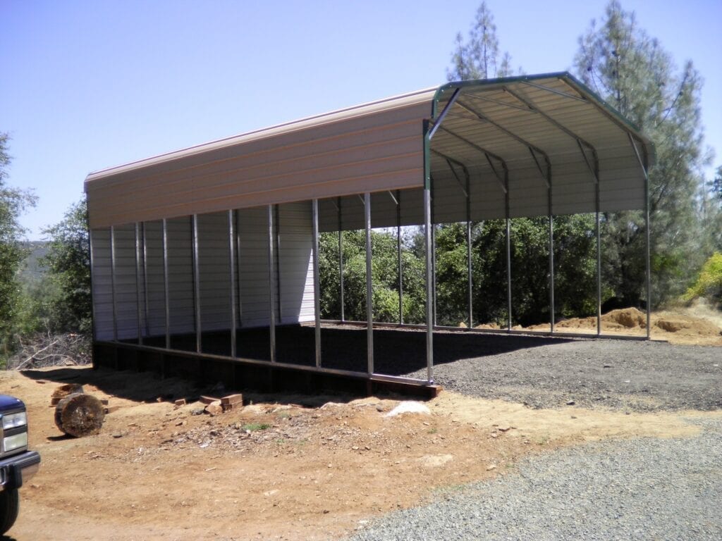 Open-sided metal carport with a brown roof on a gravel lot, surrounded by trees
