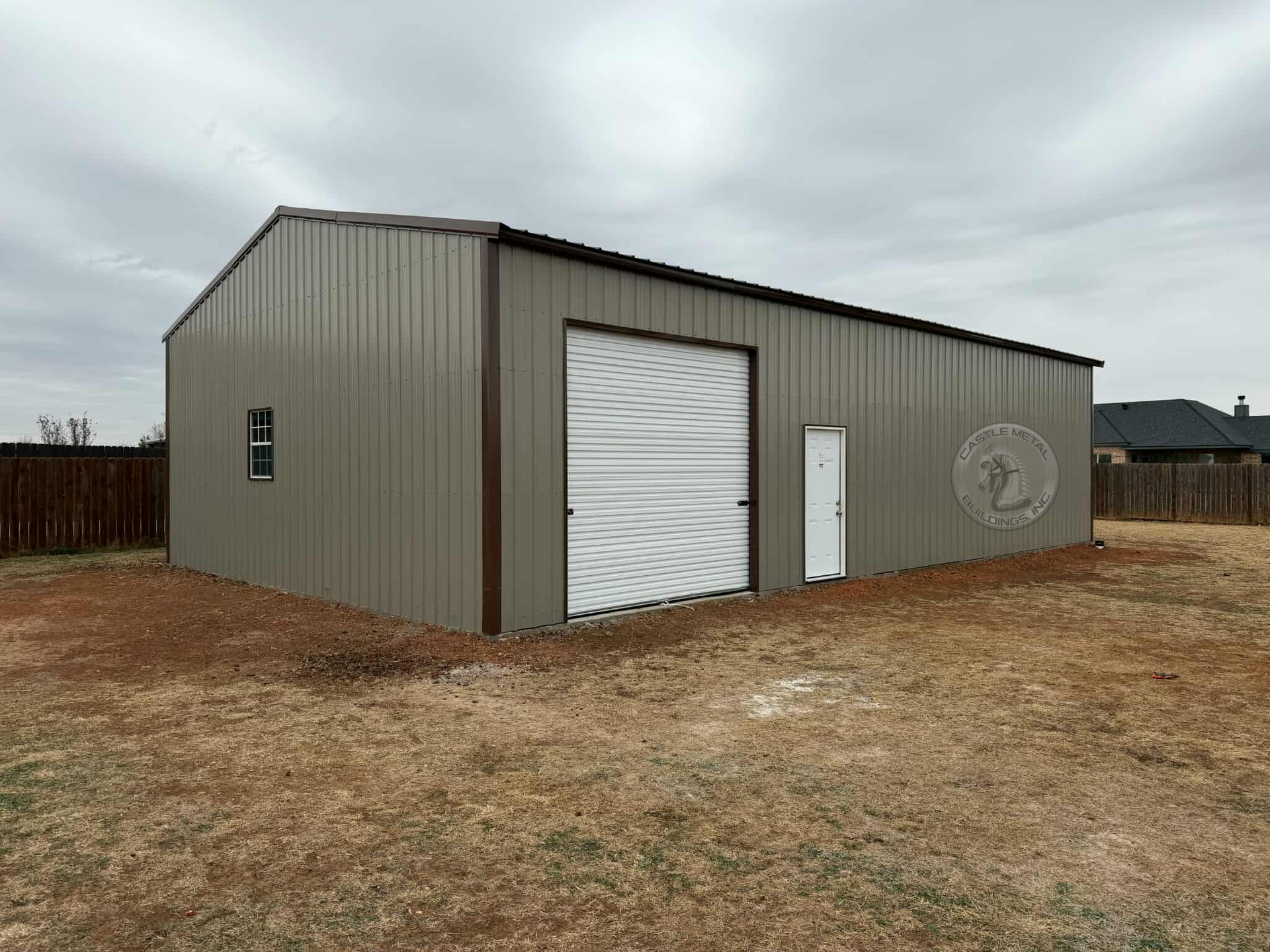 Tan metal garage building with white roll-up door on a cloudy, bare lot