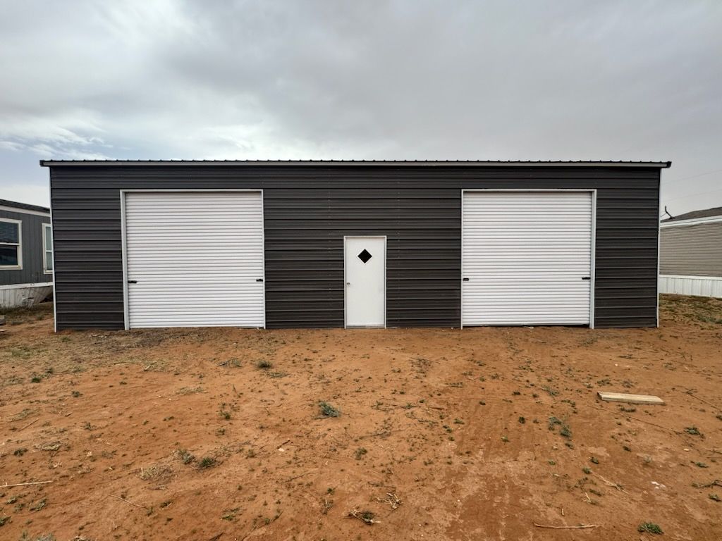 Dark metal building with two white garage doors and a central white entry door on a dirt lot under cloudy sky
