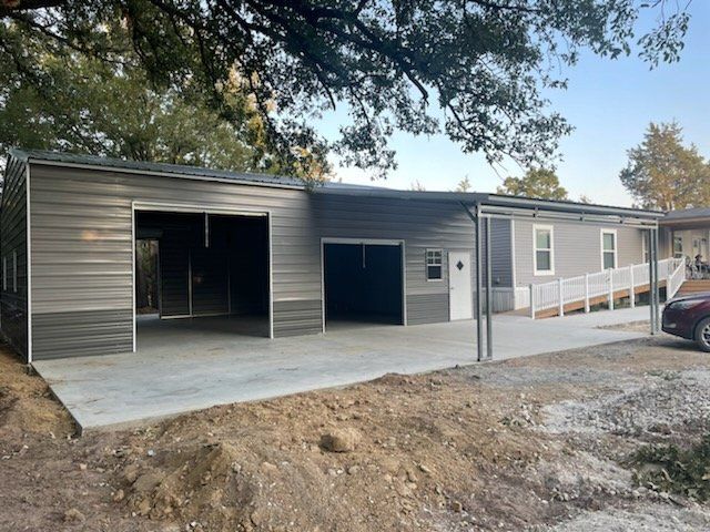 New metal building with open bays and gray siding, fronted by concrete and dirt work under trees